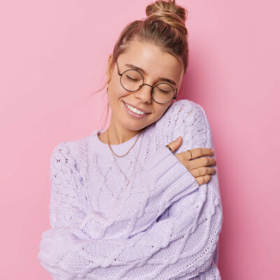 Beautiful pleased young woman with fair hair gathered in bun embraces own body wears knitted sweater expresses self love takes care of herself isolated over pink background. Self acceptance.
